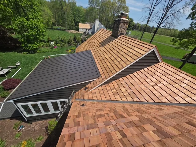 Roofing installation featuring a blend of cedar shake and metal roofing, showcasing durability and aesthetic appeal, surrounded by lush greenery in a residential setting.
