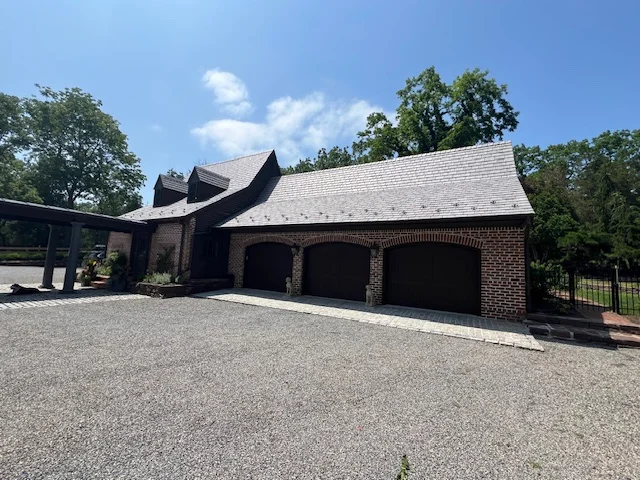 Brick home with a slate roof and three garage doors, surrounded by trees and a gravel driveway, emphasizing quality roofing services in Pipersville, PA.