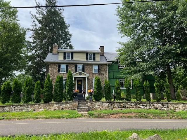 Historic stone house with a green side extension, surrounded by manicured shrubs and trees, located in a rural Bucks County setting, reflecting local architectural styles relevant to Franco Roofing Inc.'s service area.